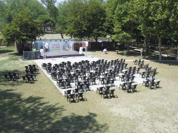 Spazio all'aperto con sedie e palco, circondato da alberi.