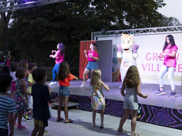 Bambini ballano davanti a un palco con animatori e una mascotte.