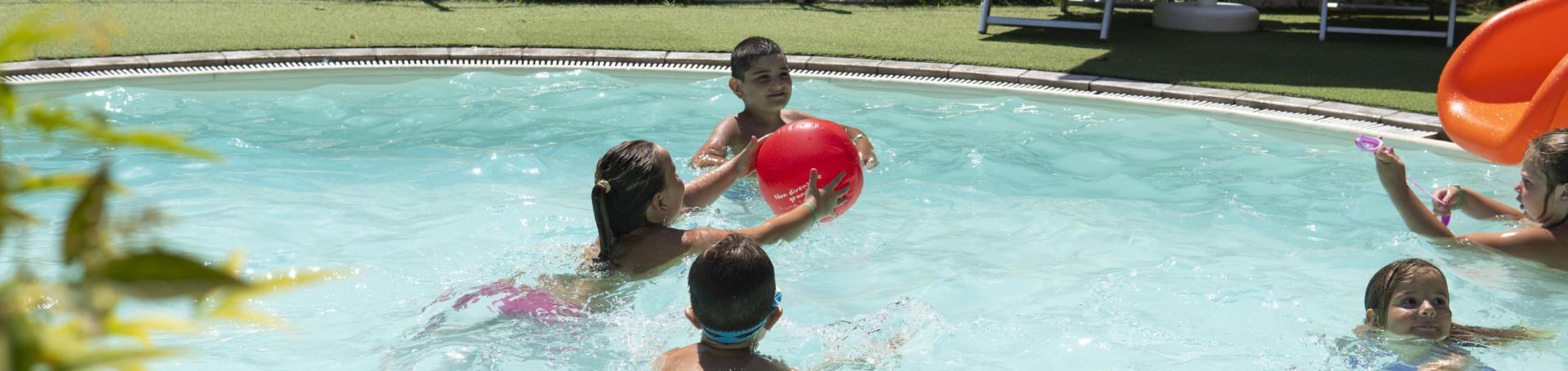 Bambini giocano in piscina con una palla rossa in una giornata soleggiata.